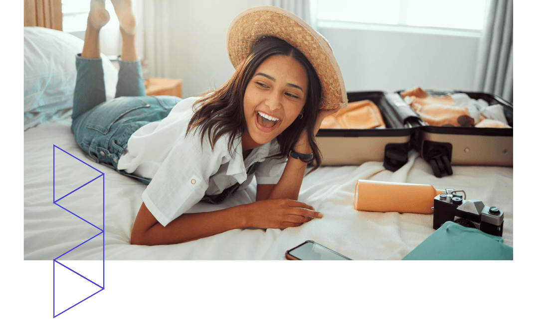 A smiling young woman in a straw hat lying on a hotel bed next to an open suitcase. 