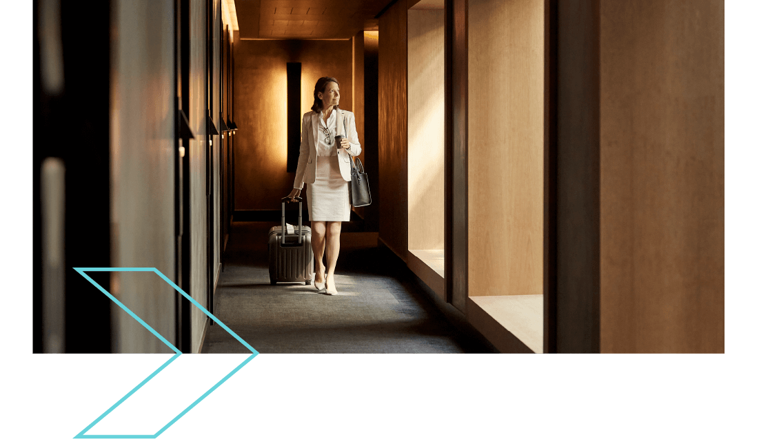 A professionally dressed woman pulling a carry on case along a hotel corridor. 