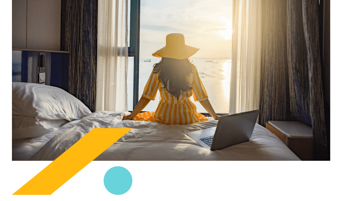Woman in a sunhat sitting on a hotel bed, looking at the sea out of an open window. 