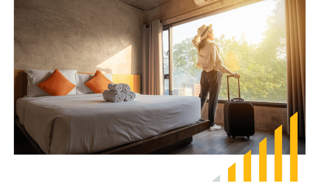 Woman with a suitcase in a hotel bedroom looking out of a sunny window with trees outside. 