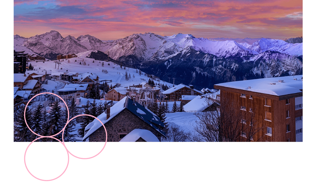 A view of snowy mountains at sunset with buildings in the foreground. 