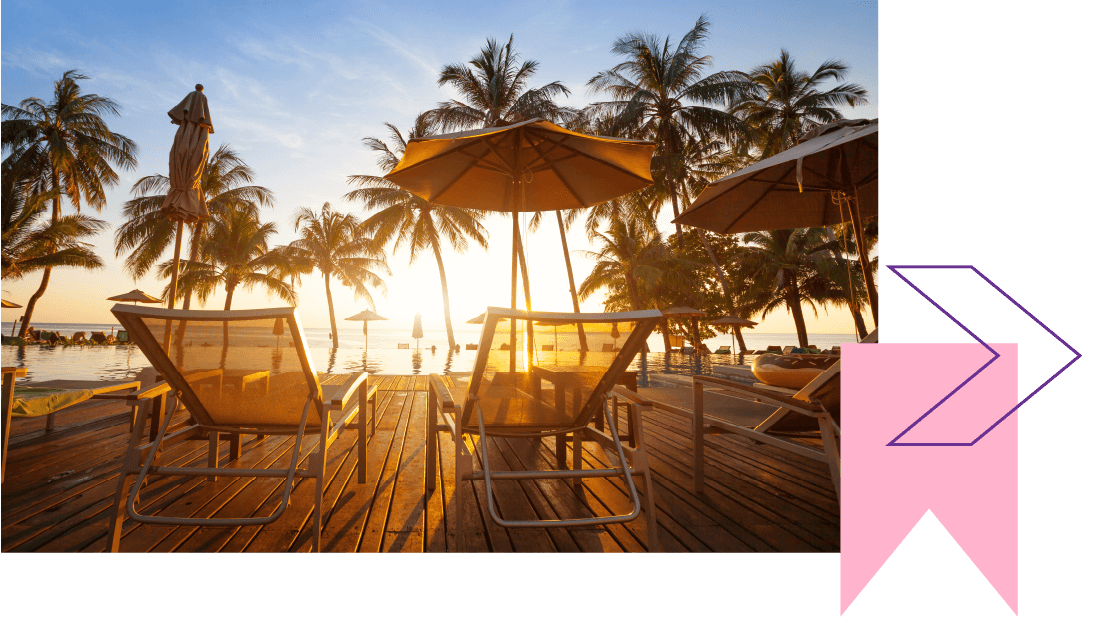 Two empty sun loungers on decking with parasols and palm trees, overlooking a beach at sunset. 