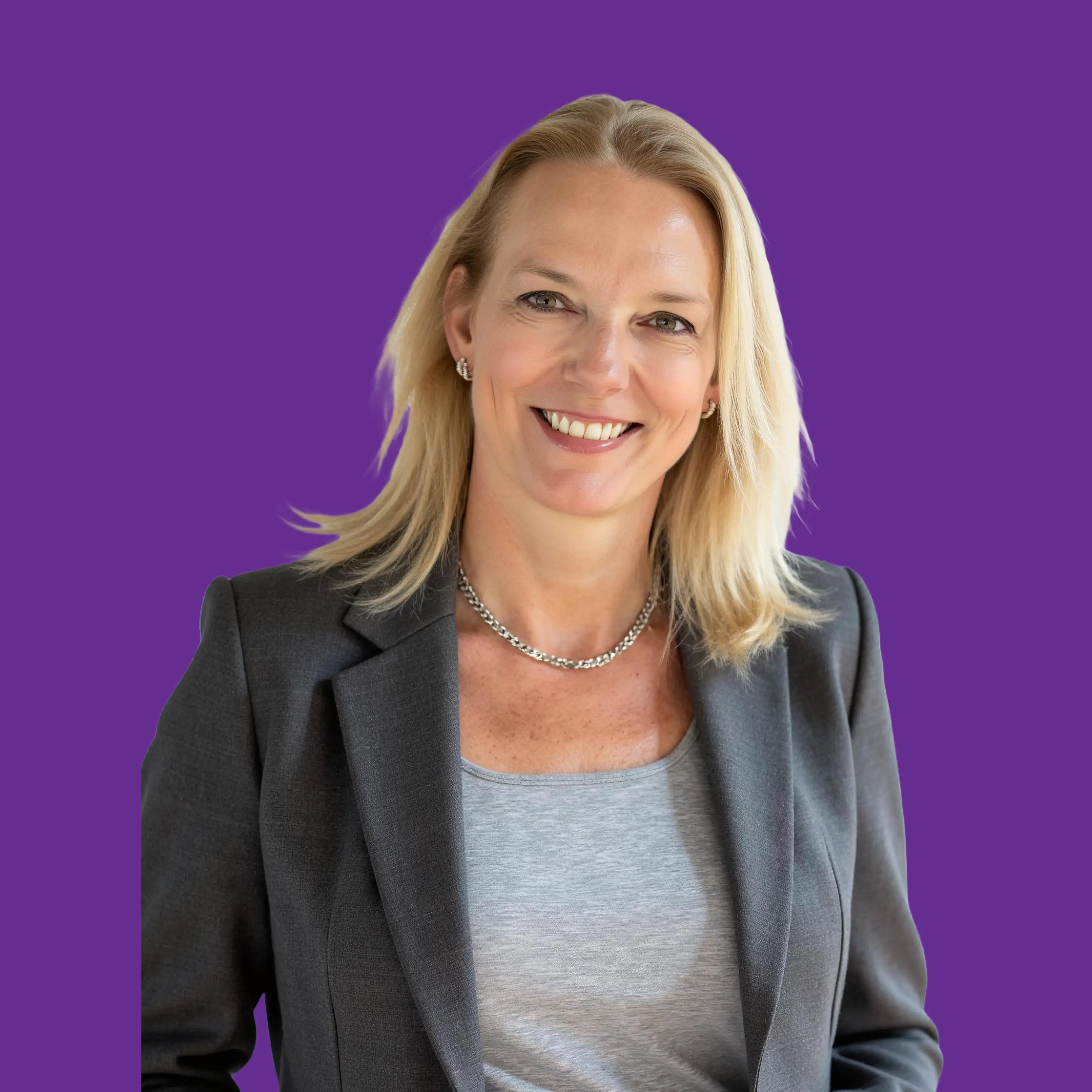 A smiling lady with short blond hair, silver earrings and necklace, wearing a grey shirt and blazer.