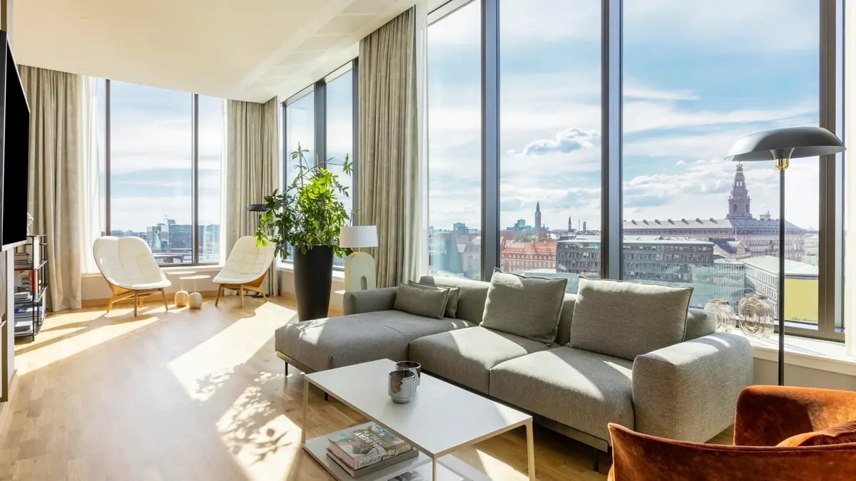 A sunny room with grey a gray coach, plants, white chairs, and a table with vast windows and a city line in the background.