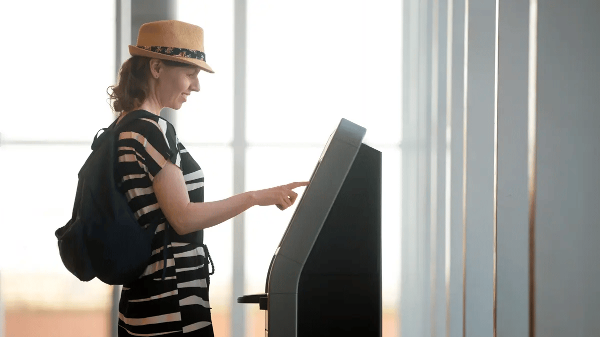 A woman with black and white dress, a backpack and a hat, clicking on a screen at an airport.