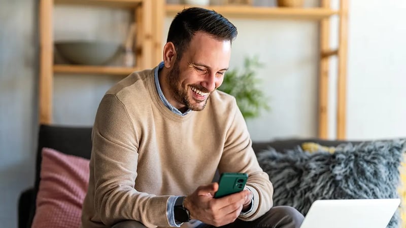Uomo che tiene un telefono nella mano destra, guarda un computer portatile e sorride.