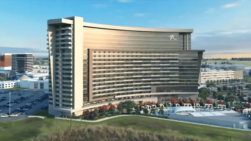 A large greyish hotel in a wave-like shape with a green area and a swimming pool at the front, and parking lots in the back.