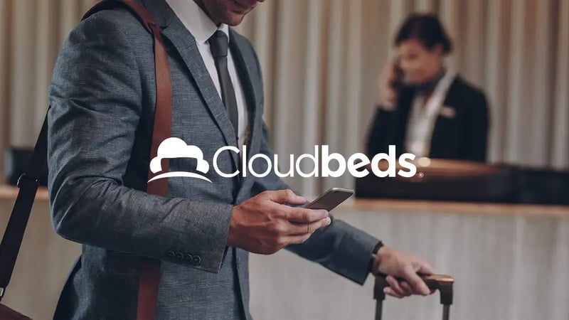 Un uomo in giacca e cravatta con il telefono in mano e una valigia alla scrivania di un hotel con il logo Cloudbeds in bianco in sovrimpressione.