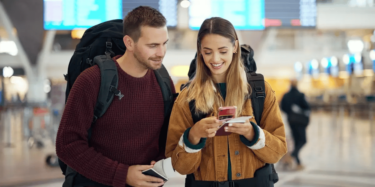 Two people with backpacks hold their passports at the airport and are looking down at their phone, smiling.
