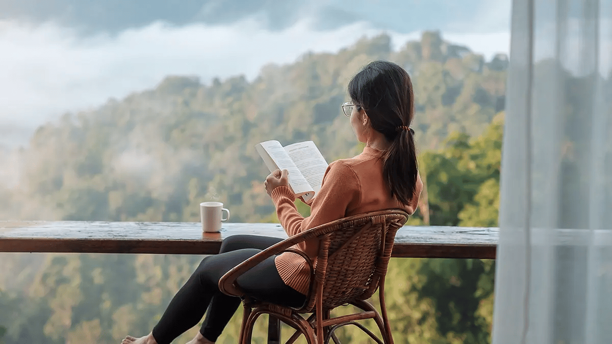 View from behind of a person sat in a wicker chair reading a book, with tree-covered mountains in the background. 