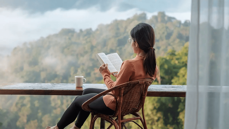 View from behind of a person sat in a wicker chair reading a book, with tree-covered mountains in the background. 