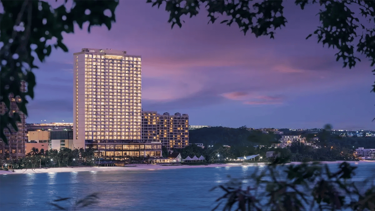 A photo of Dusit's iconic hotel at the seaside, taken at dusk with a frame of dark branches and leaves at the front.