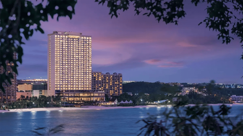 A photo of Dusit's iconic hotel at the seaside, taken at dusk with a frame of dark branches and leaves at the front.