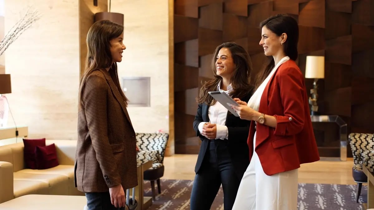 Three people chatting in a hotel's lobby, wearing smart clothes with one of them holding an iPad and taking notes.