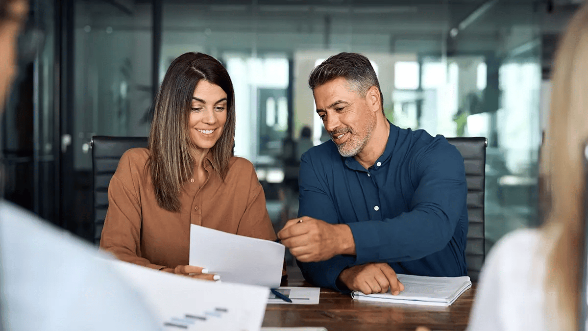 A woman and a man sat at a desk looking at a piece of paper and smiling.