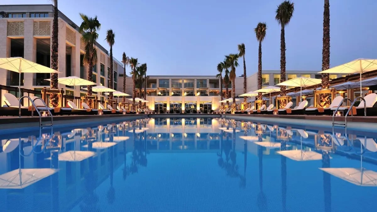 A hotel's pool at dusk with sunbeds and umbrellas and their reflections in the water, surrounded by two-floor white hotel with Arabic patterns.