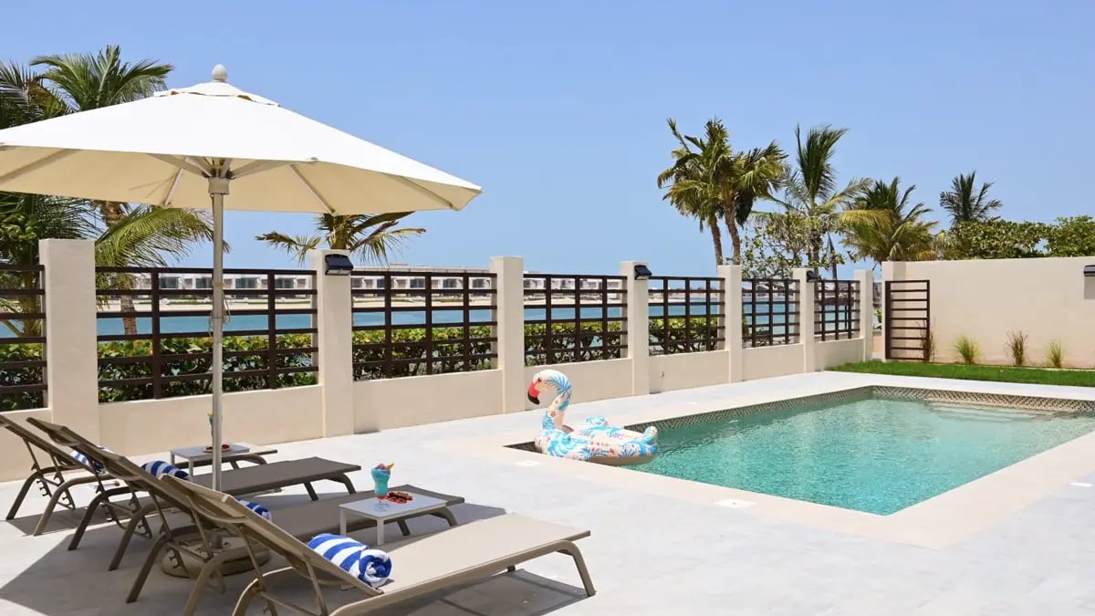 A small swimming pool with 3 sunbeds under an umbrella with the sea and palms in the background.