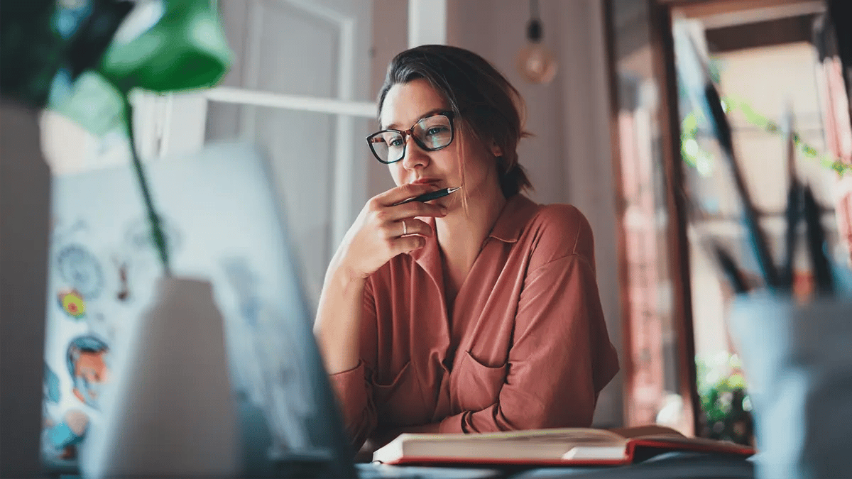 A lady with glasses, seating at a desk with a pen in hand, looking at her laptop's screen, ready to take notes in the book opened in front of her.
