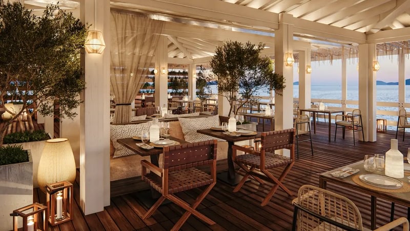 A panoramic photo of a covered outdoors' restaurant with wooden tables and chairs overlooking the sea.