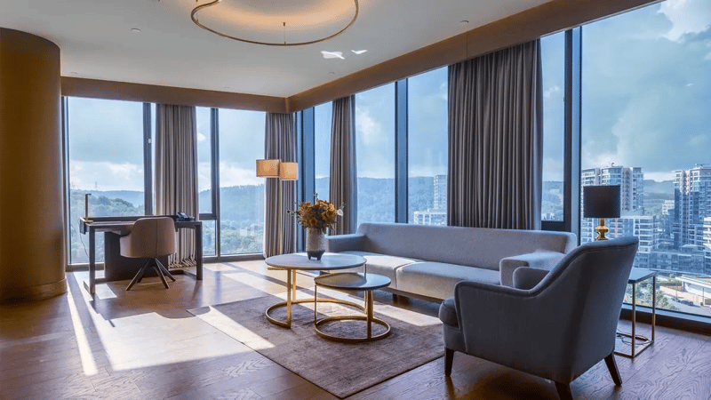 A sunny hotel room on an upper floor with a table and a few coaches and sofas and a 360 degree city view through French windows.