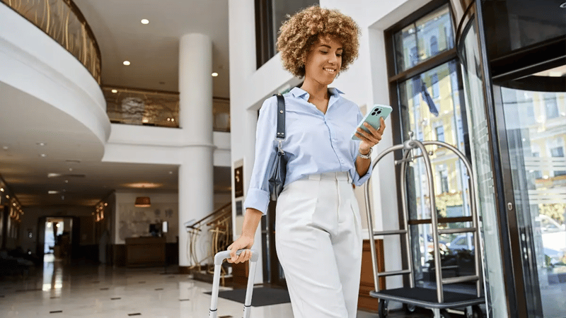 A smiling lady walking through a hotel's lobby with her suitcase while looking at her phone.