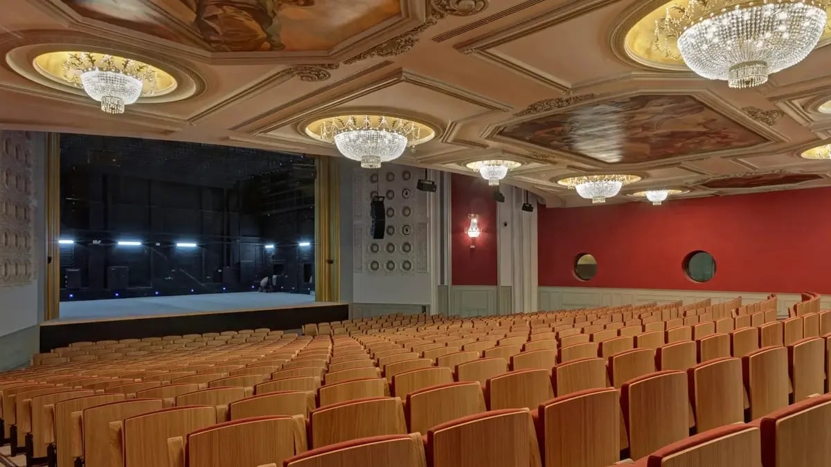 A hotel's theatre with red walls, beautiful golden chandeliers and rows of wooden chairs leading towards the stage.