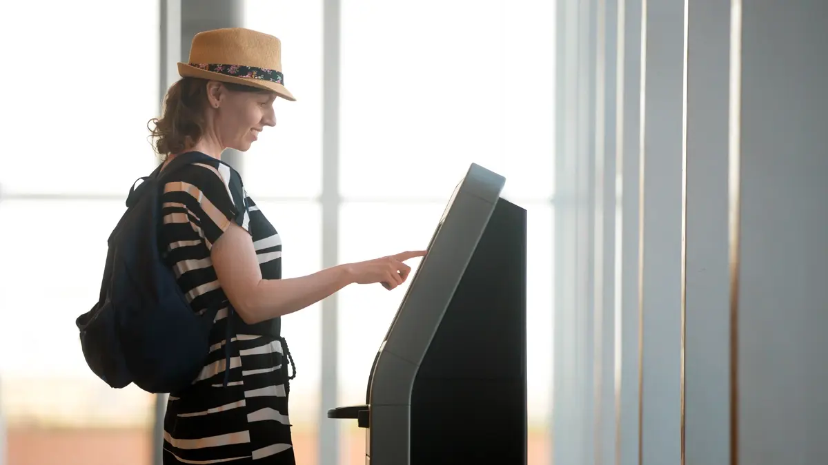 A woman with black and white dress, a backpack and a hat, clicking on a screen at an airport.