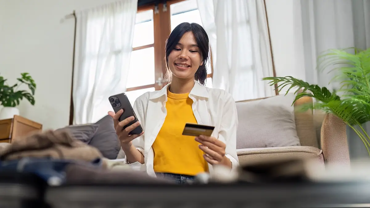 Donna con in mano una carta di credito e un telefono, con una valigia sfocata in primo piano.