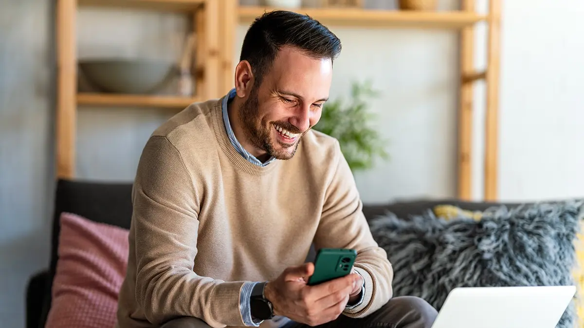 Uomo che tiene un telefono nella mano destra, guarda un computer portatile e sorride.