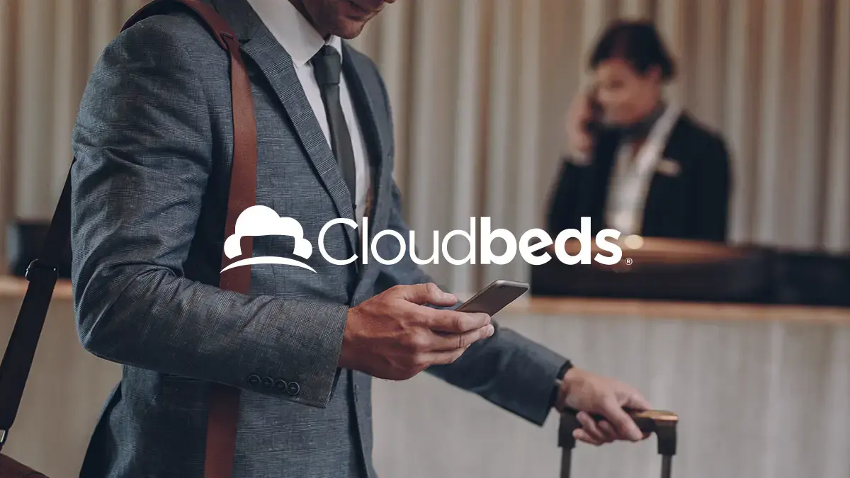 Ein Hauptdarsteller im Anzug hält sein Telefon und einen Koffer an einem Hotelschreibtisch mit dem Cloudbeds-Logo in Weiß als Overlay.