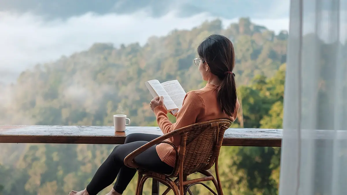 L'immagine raffigura una persona seduta a un tavolo di legno, che legge un libro. Si trova in un ambiente accogliente e naturale con una vista panoramica di montagne o colline sullo sfondo, forse avvolte nella nebbia. La persona indossa un maglione e degli occhiali, e c'è una tazza appoggiata sul tavolo. L'atmosfera generale appare tranquilla e invitante, suggerendo un momento di relax e contemplazione.