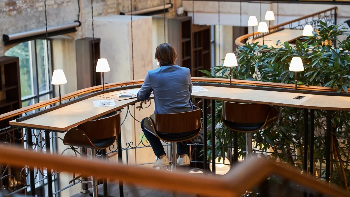 A man sitting on the middle out of three chairs at a corner long desk overlooking the lower floor in a hotel lobby.