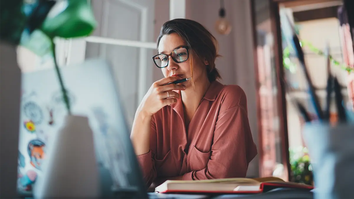 A lady with glasses, seating at a desk with a pen in hand, looking at her laptop's screen, ready to take notes in the book opened in front of her.