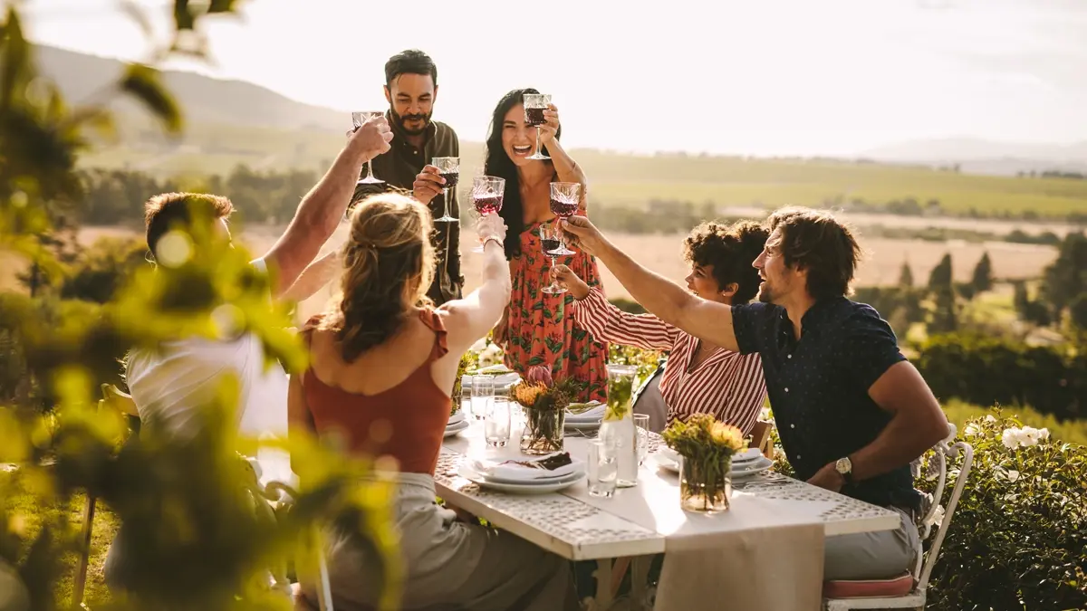 A group of friends wearing summery clothes, sitting at a table in a garden making a toast.